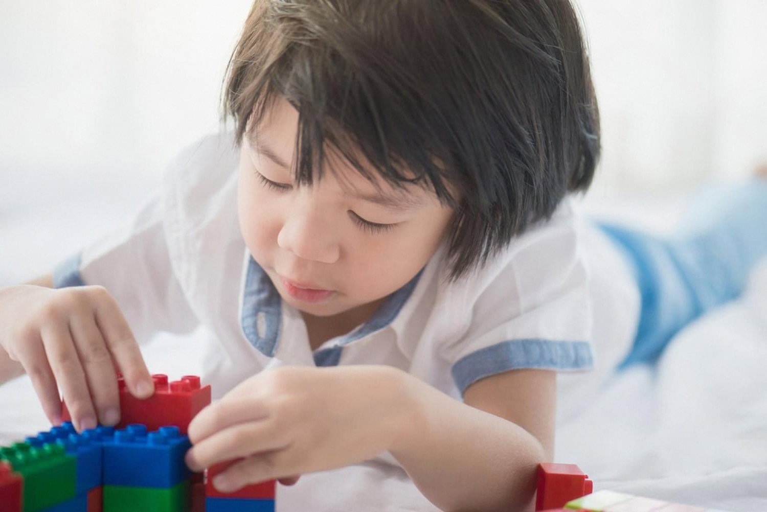 Young boy focused on assembling colourful plastic building blocks on a bed